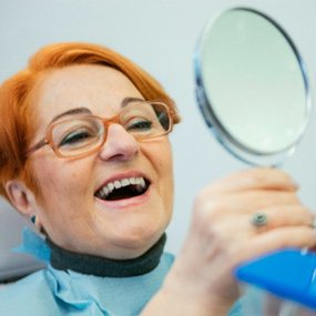 An older smiling woman admiring her dentures in a hand mirror